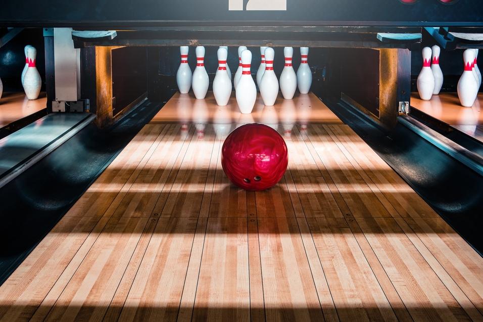 Ein roter Bowlingball rollt auf einer Holzbahn in Richtung weißer Kegel mit roten Ringen im Hintergrund.