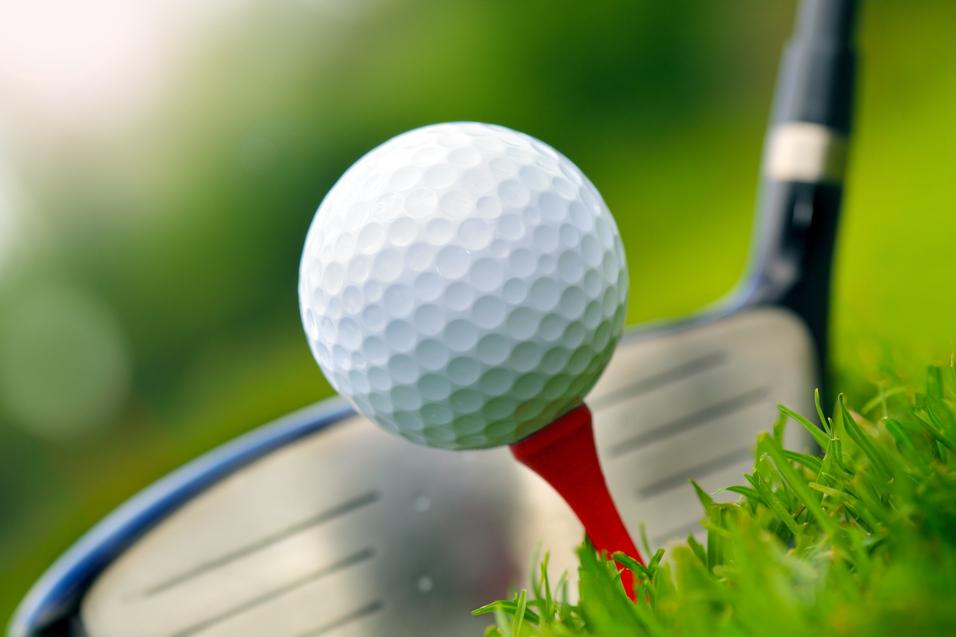 Nahaufnahme eines Golfballs auf einem roten Tee, mit einem Golfschläger im Hintergrund auf grünem Gras.