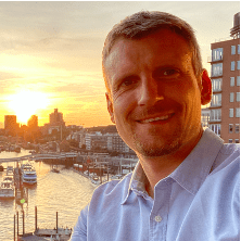 Mann in hellblauer Hemd sitzt am Hafen, Sonnenuntergang im Hintergrund, Boote im Wasser sichtbar.